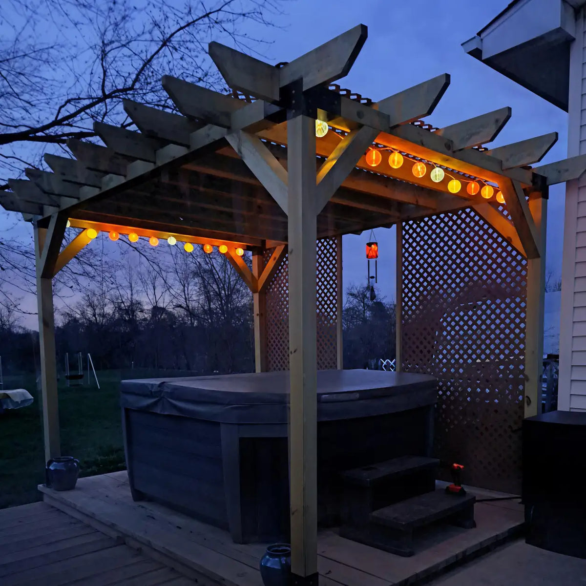 Angled view of a wooden pergola over an outdoor hot tub at dusk, featuring black metal brackets, glowing string lights and lattice privacy screens.