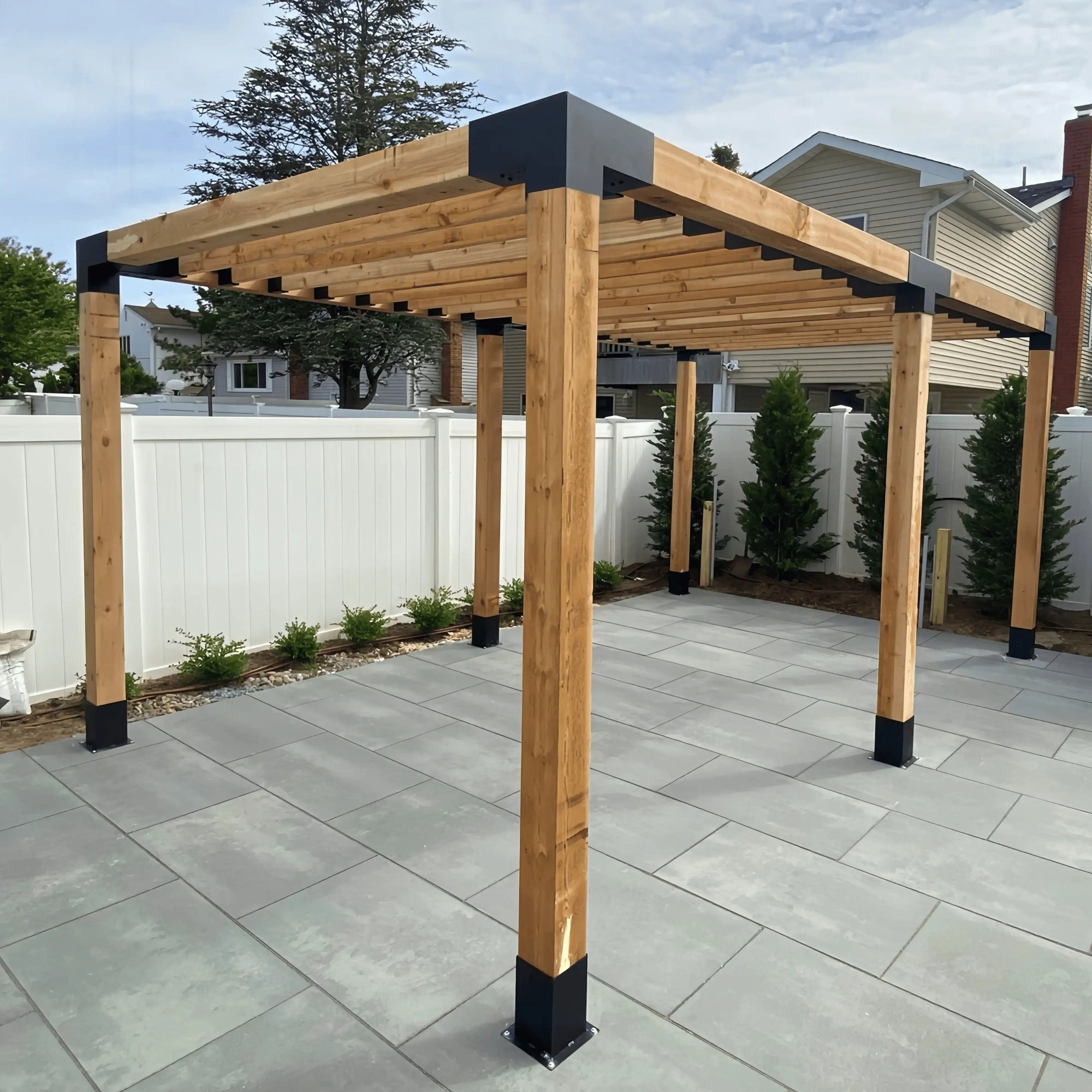 Wooden pergola kit installed in a backyard, featuring Jurrque brackets, placed on a flat tiled surface with surrounding greenery and a white fence.