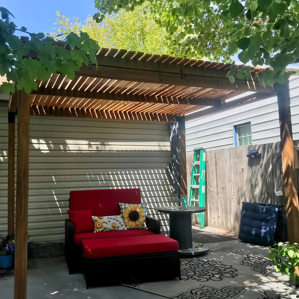 Angled view of a wooden pergola with a slatted roof shading a patio seating area with a red cushioned loveseat, ottoman and side table.